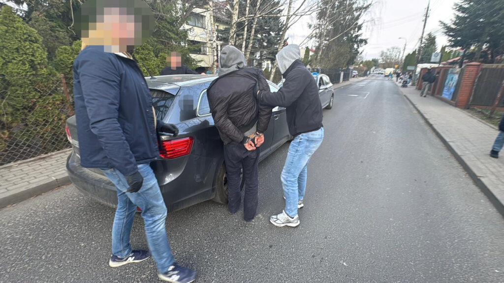 Mężczyzna w szarym kapturze jest zatrzymywany przez dwóch policjantów na ulicy, obok samochodu osobowego.