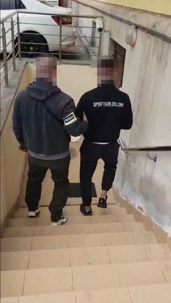 Policjant prowadzi mężczyznę w dresie po schodach w kierunku wyjścia z budynku.