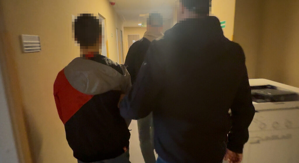 Mężczyzna w kurtce sportowej prowadzony przez dwóch funkcjonariuszy w korytarzu budynku.