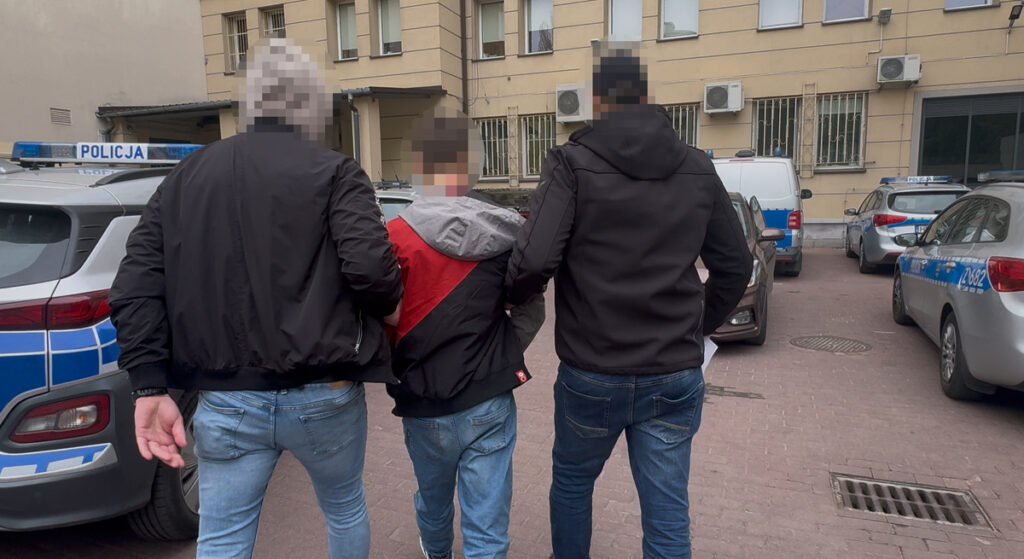 Mężczyźni prowadzą młodego chłopca w kierunku radiowozów policyjnych na podwórzu budynku.