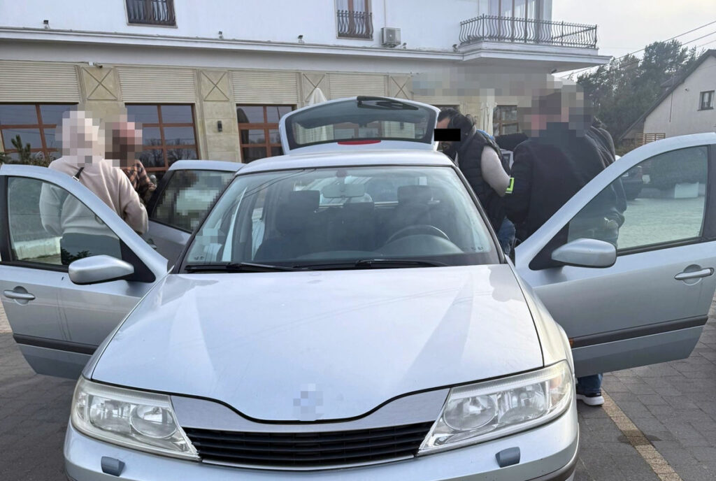 Grupa osób rozmawia przy otwartym bagażniku srebrnego samochodu na parkingu przed budynkiem.