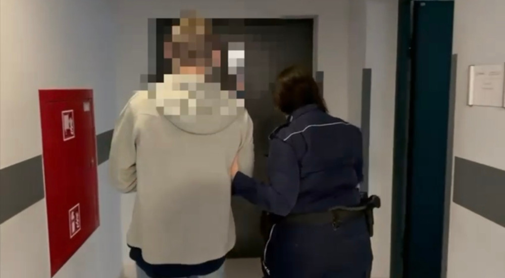 Mężczyzna w szarym ubraniu prowadzony przez funkcjonariusza policji w korytarzu budynku.