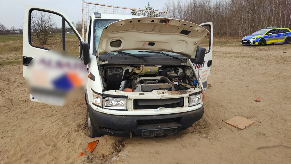Otwarty samochód dostawczy z widocznym silnikiem, zaparkowany na piaszczystym terenie, obok radiowóz policyjny.