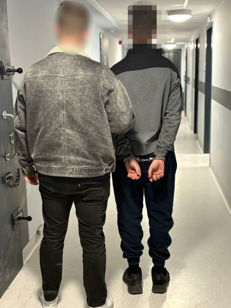 Mężczyzna w szarym dżinsowym płaszczu prowadzi osobę w kajdankach przez korytarz w budynku.