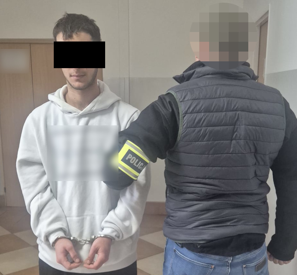 Mężczyzna w białej bluzie z kajdankami na rękach stoi w pomieszczeniu, podczas gdy policjant z oznakowaniem na rękawie go pilnuje.