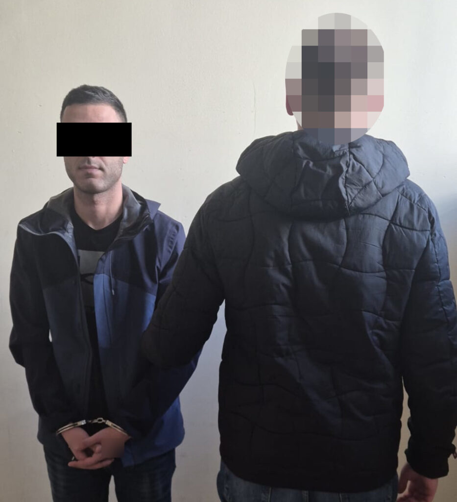 Mężczyzna w kajdankach stoi z rękami z tyłu, obok niego stoi drugi mężczyzna tyłem, w czarnej kurtce.