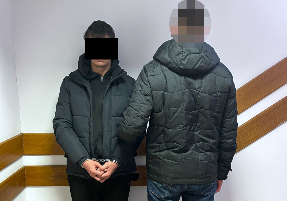 Mężczyzna w rękach policji stoi w pomieszczeniu z drewnianymi panelami, z rękami związanymi na plecach.