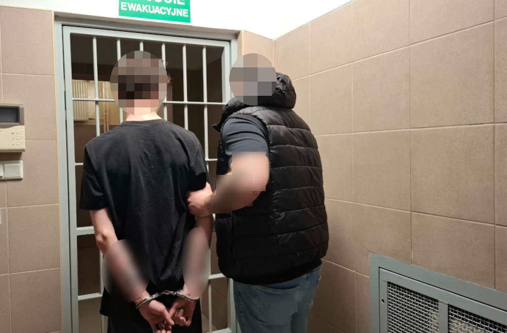 Młody mężczyzna w kajdankach prowadzony przez funkcjonariusza w pomieszczeniu z kratami.