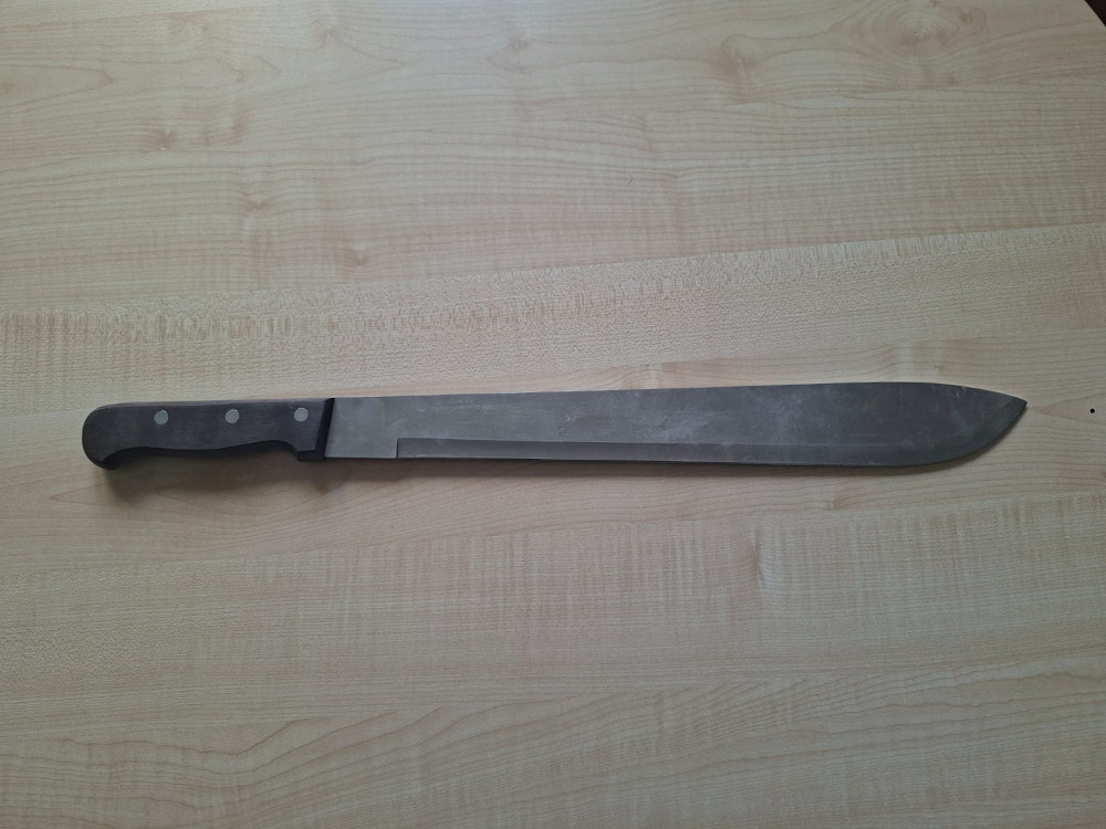 Długi nóż z metalowym ostrzem leży na drewnianym blacie. Rękojeść jest wykonana z ciemnego drewna z trzema metalowymi wstawkami.