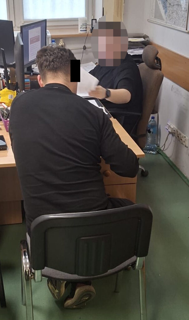 Mężczyzna w czarnym t-shircie siedzi na krześle, podczas gdy druga osoba w biurze przegląda dokumenty.