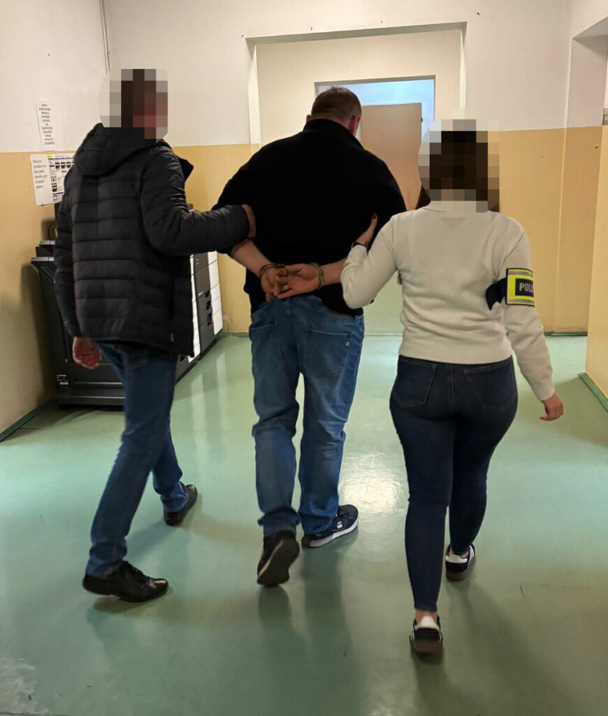 Mężczyzna w kajdankach prowadzony przez dwóch funkcjonariuszy w korytarzu policyjnym.