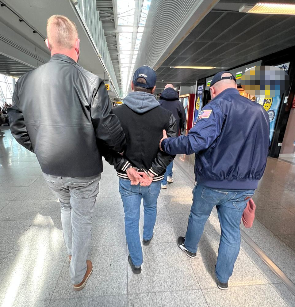 Trzech mężczyzn prowadzi zatrzymanego, który ma założone kajdanki, w korytarzu lotniska.