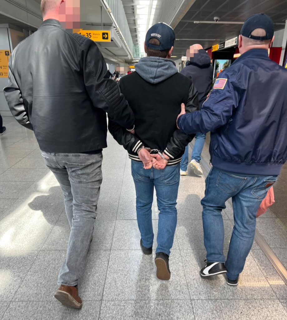 Mężczyzna w kajdankach prowadzony przez dwóch funkcjonariuszy w terminalu lotniska.