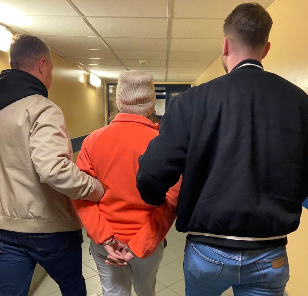 Kobieta w pomarańczowej bluzie prowadzona przez dwóch mężczyzn w korytarzu budynku, z rękami skuty w kajdankach.