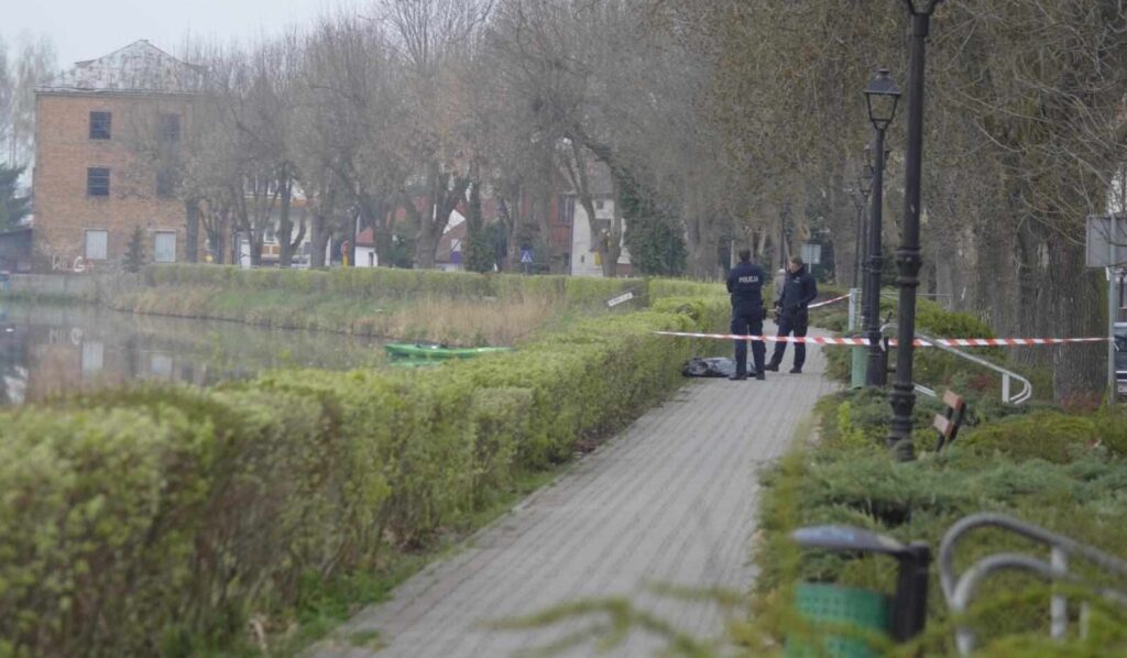Policjanci zabezpieczają teren przy chodniku wzdłuż rzeki, obok zielonego żywopłotu.