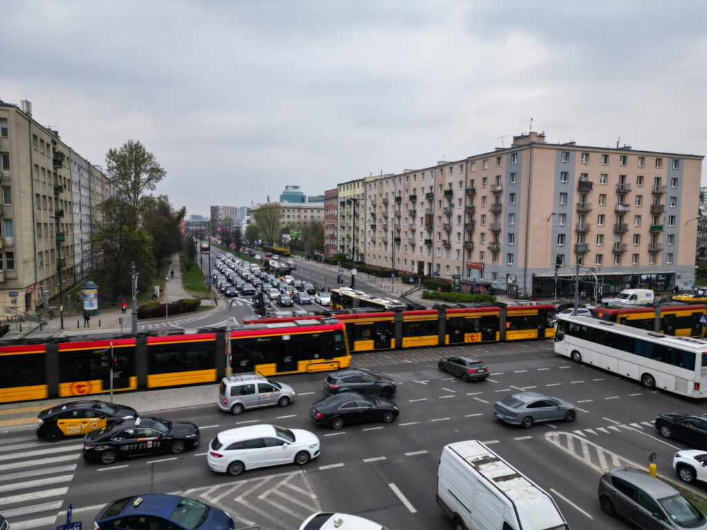 Ruch tramwajowy przejeżdża przez zatłoczoną ulicę z samochodami, obok bloków mieszkalnych i biurowców.