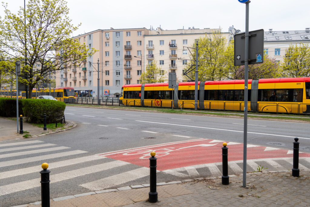 Zielony tramwaj z czerwonym dachem przejeżdża obok nowoczesnych budynków mieszkalnych, w otoczeniu drzew.