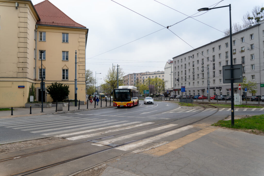 Autobus komunikacji miejskiej skręca w prawo na skrzyżowaniu, obok nowoczesnych budynków mieszkalnych. W tle przechodnie korzystają z przejścia dla pieszych.