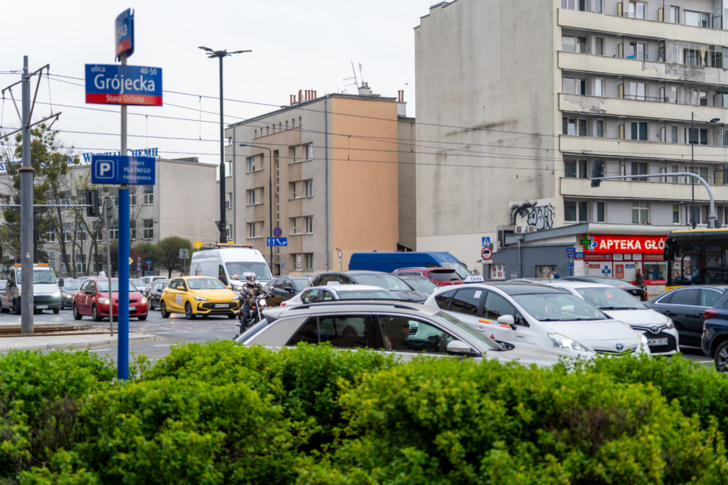 Ruchliwa ulica z zatorami drogowymi, w tle widoczny budynek z apteką i znak ulicy Grójeckiej.