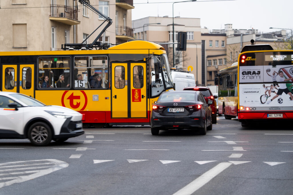 Tramwaj w żółto-czarnych barwach zatrzymany na skrzyżowaniu, obok przejeżdżające samochody osobowe w ruchu miejskim.