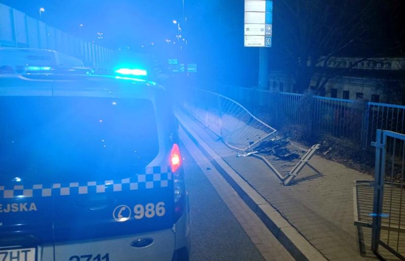 Policyjny radiowóz z włączonymi sygnałami świetlnymi stoi przy uszkodzonej barierce na drodze nocą.