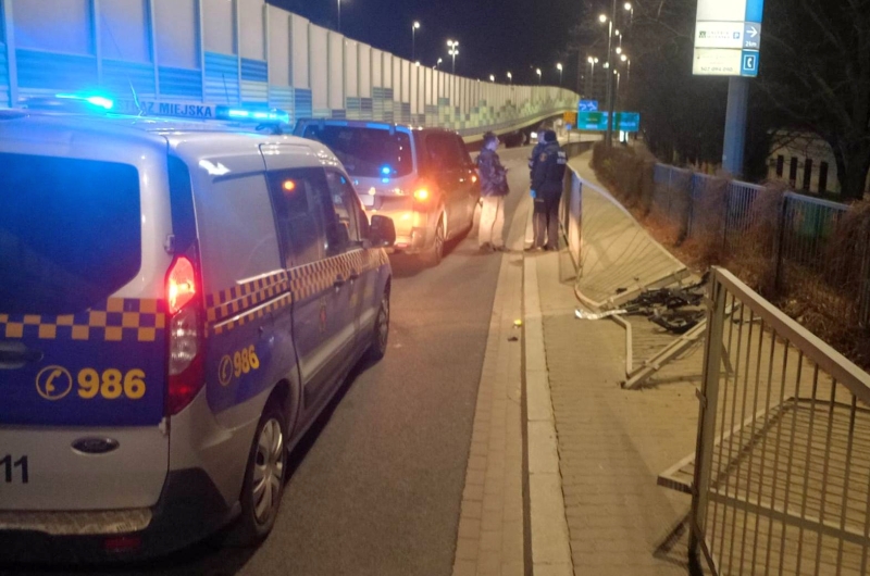 Na drodze stoi radiowóz z włączonymi sygnałami świetlnymi, a obok rozmawia kilka osób.