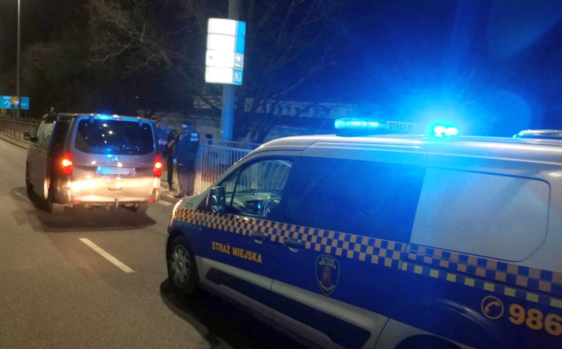 Samochód straży miejskiej z włączonymi sygnałami świetlnymi zatrzymany przy ulicy, obok inny pojazd.