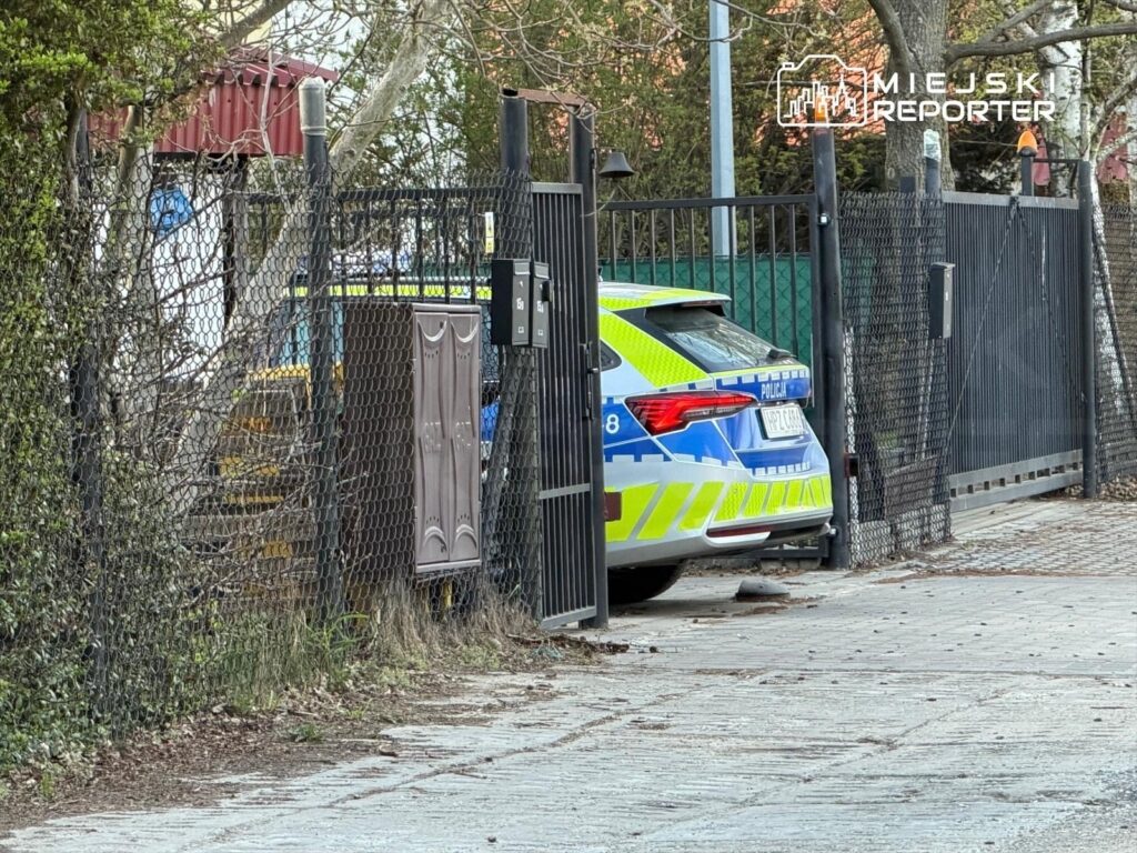Policyjny radiowóz zaparkowany przy zamkniętej bramie w ogrodzeniu, w tle widoczne drzewa i schody.