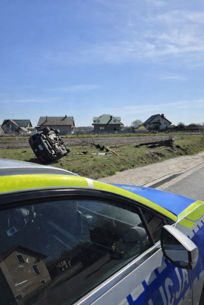 Przewrócony samochód leży na boku w pobliżu drogi, obok widoczne domy i puste pola. W tle niebo bezchmurne.