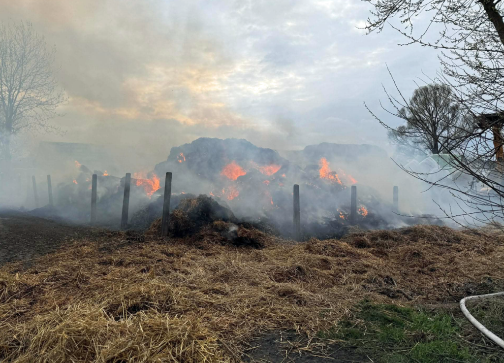 Płonąca sterta siana otoczona dymem, w tle widać zarysy drzew i niebo.