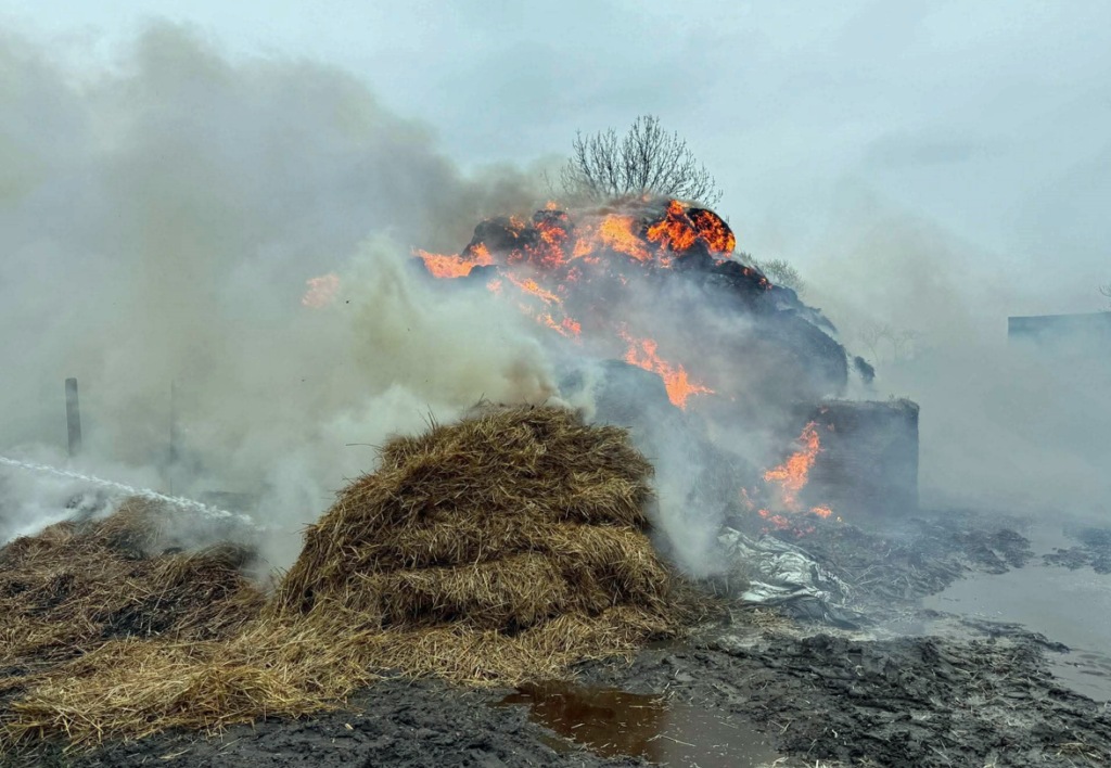 W tle płonąca sterta siana, otoczona gęstym dymem, w mokrym terenie.