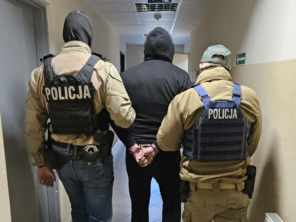 Dwóch policjantów w mundurach prowadzi zatrzymanego mężczyznę w czarnej bluzie przez korytarz budynku.