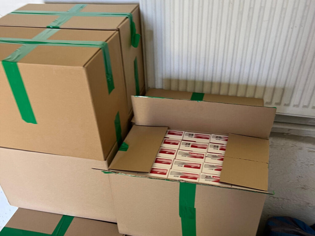 Kartony z zieloną taśmą, w tym jeden otwarty z paczkami, stoją na podłodze w pomieszczeniu z białą ścianą.