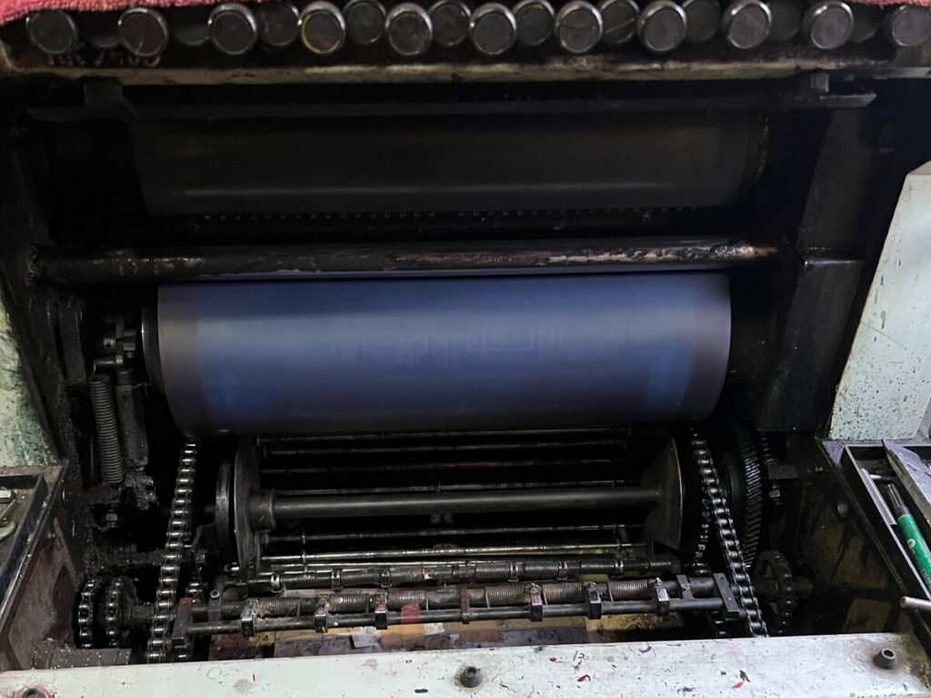 Maszyna drukarska z obrotowym cylindrem i zębatkami w ciemnym otoczeniu roboczym.