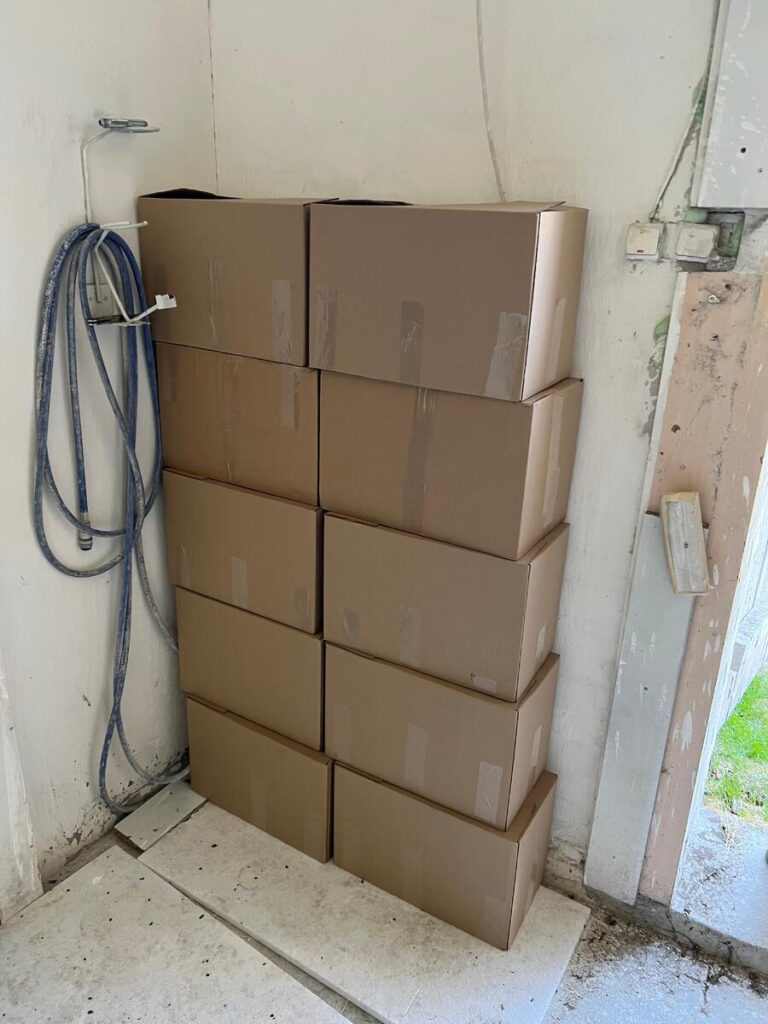 Dziesięć kartonów ustawionych w pionie w rogu pomieszczenia, obok węża i ściany z nierówną powierzchnią.