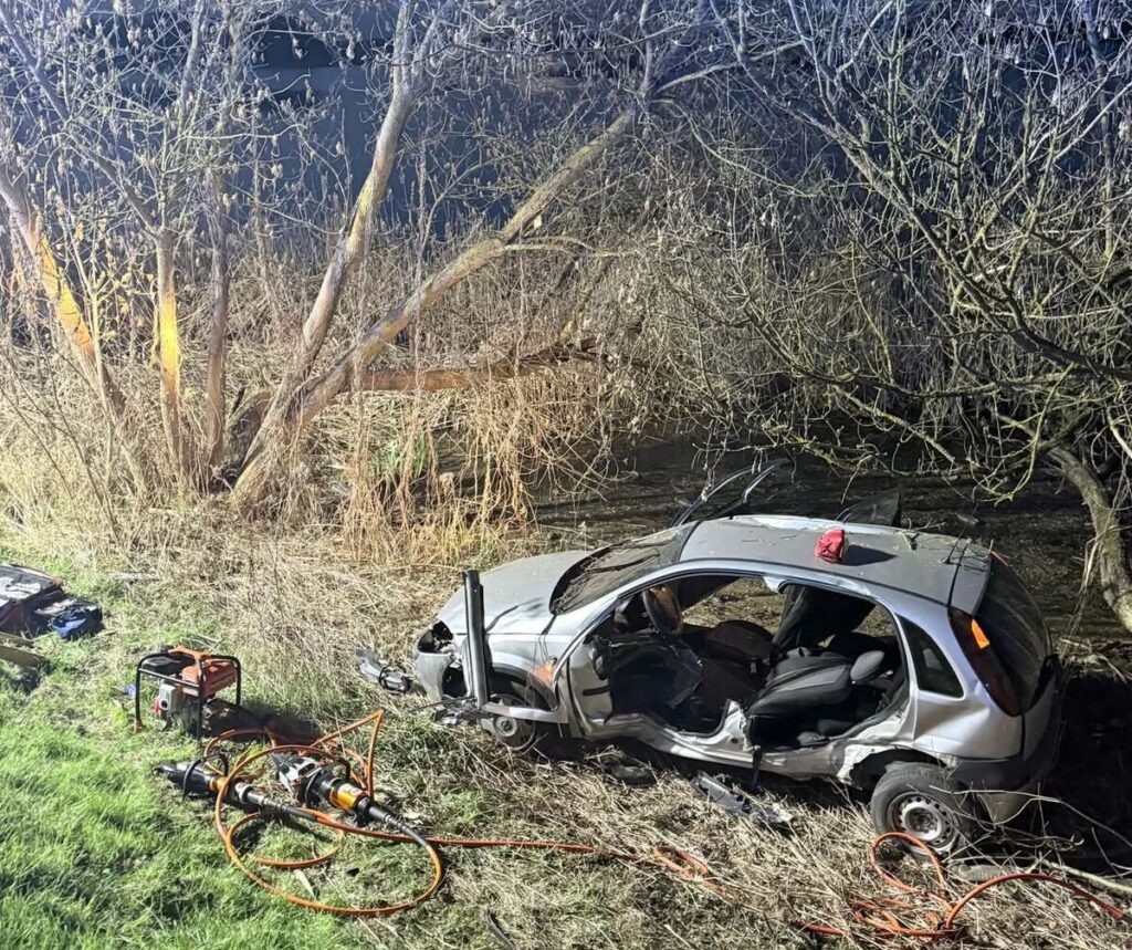 Uszkodzony samochód osobowy stoi na poboczu, otoczony przez gęste krzewy, z narzędziami i sprzętem ratunkowym obok.