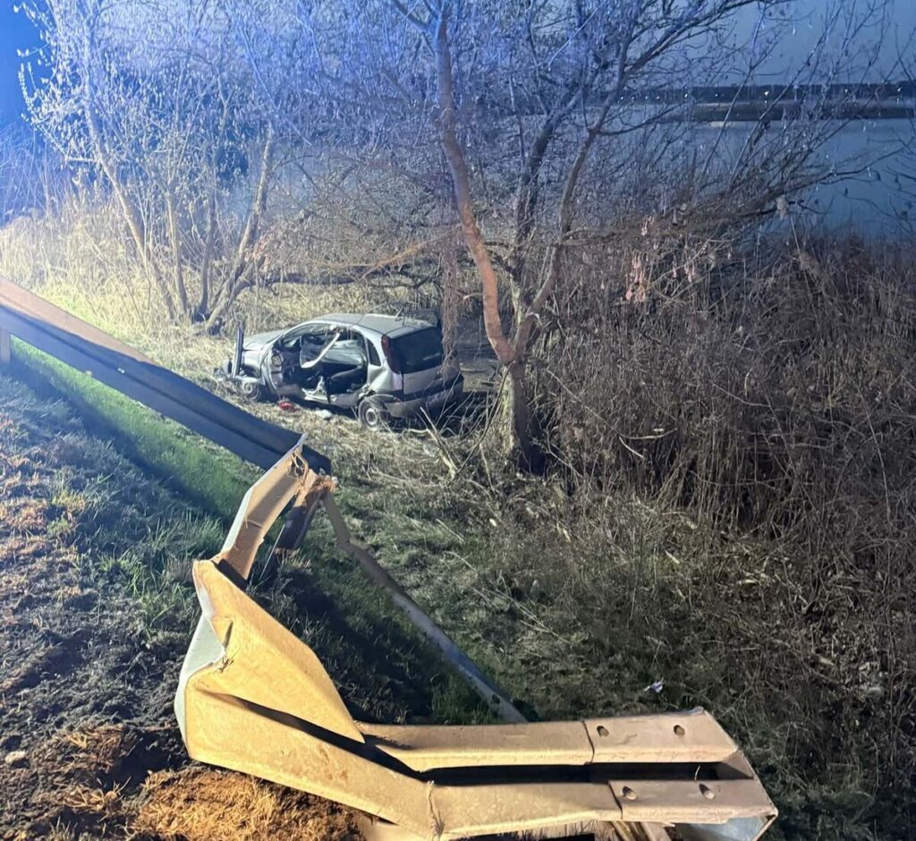 Zniszczony samochód osobowy stoi w zaroślach obok drogi, z wygiętą barierką ochronną w pierwszym planie.