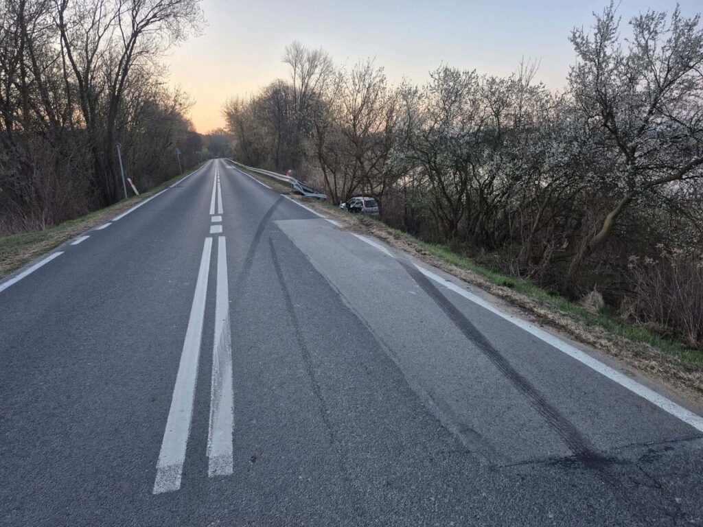 Pusta droga z widocznymi śladami opon, w tle drzewa i porzucony pojazd w zaroślach.