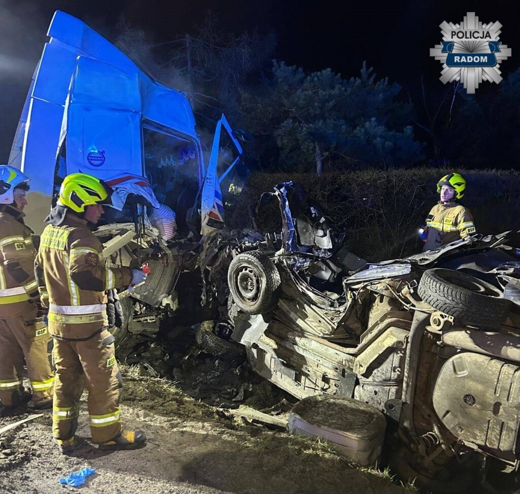 Strażacy pracują nad wypadkiem drogowym, gdzie ciężarówka zderzyła się z osobowym samochodem w ciemnym otoczeniu.