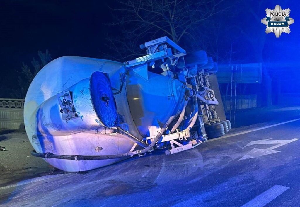 Cementowóz leży na boku na drodze, otoczony niebieskim światłem policyjnym w nocy.