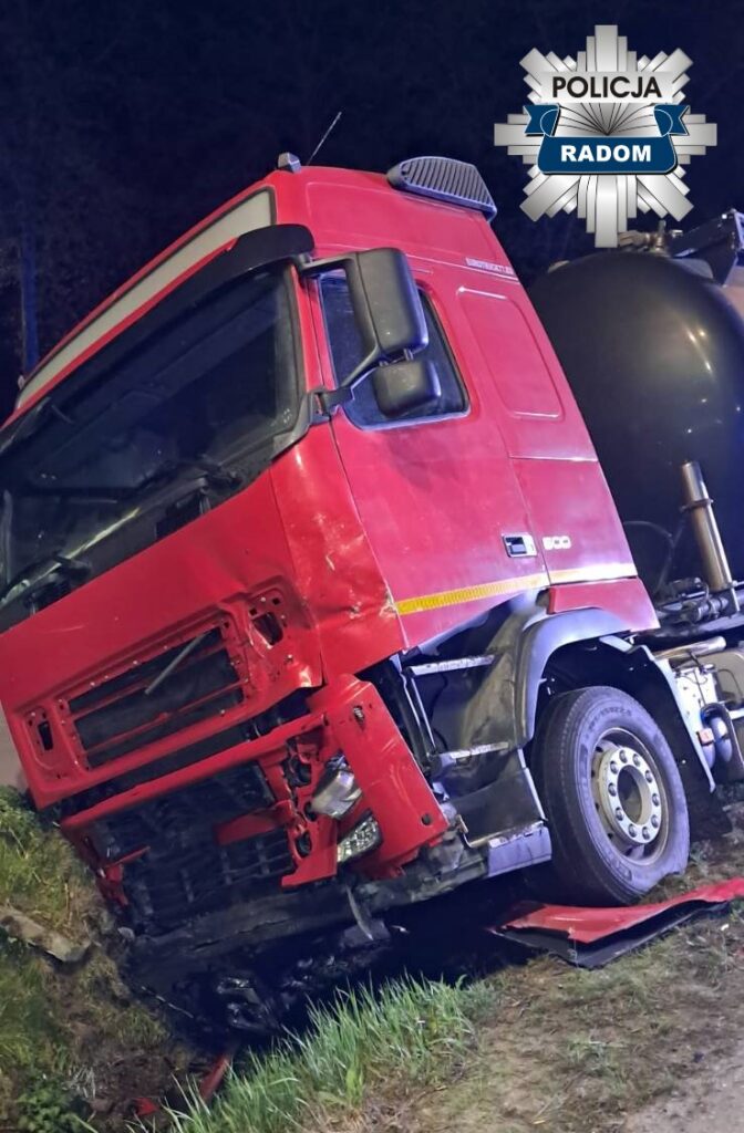 Czerwony ciężarówka leży na boku w rowie, z uszkodzoną przednią częścią, otoczona trawą i drzewami.