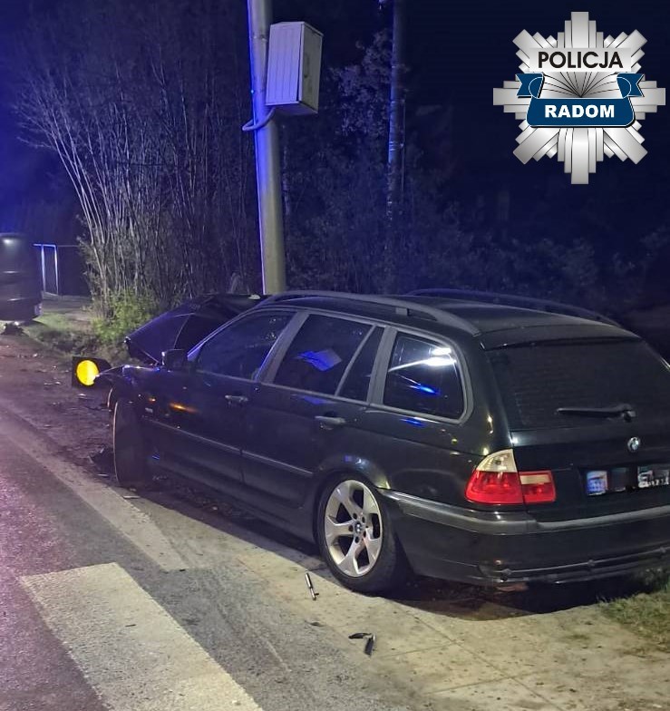 Zielony samochód osobowy zaparkowany na poboczu drogi, obok widoczne uszkodzenia i niebieskie światła policyjne.