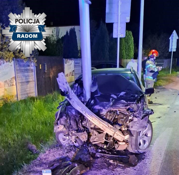 Uszkodzony samochód osobowy zderzył się z słupem na poboczu drogi, obok stoi ratownik w kasku.