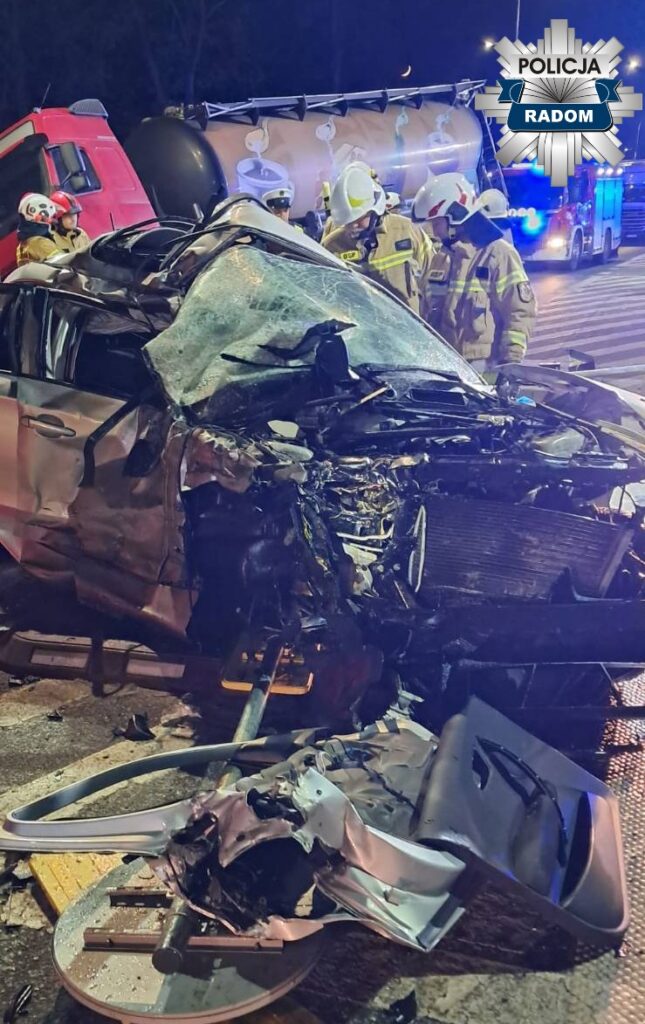 Rozbite auto osobowe z uszkodzoną przednią częścią, w otoczeniu strażaków i pojazdów ratunkowych na drodze.