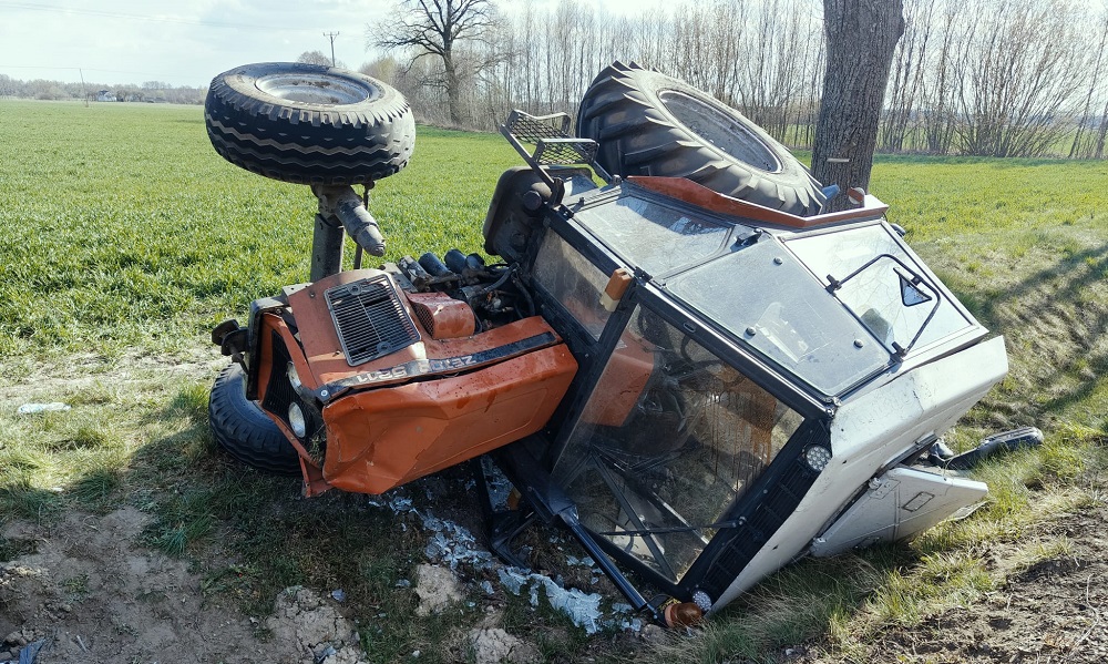 Przewrócony traktor leży na boku na polu, z jednym kołem uniesionym w górę, otoczony zieloną trawą.