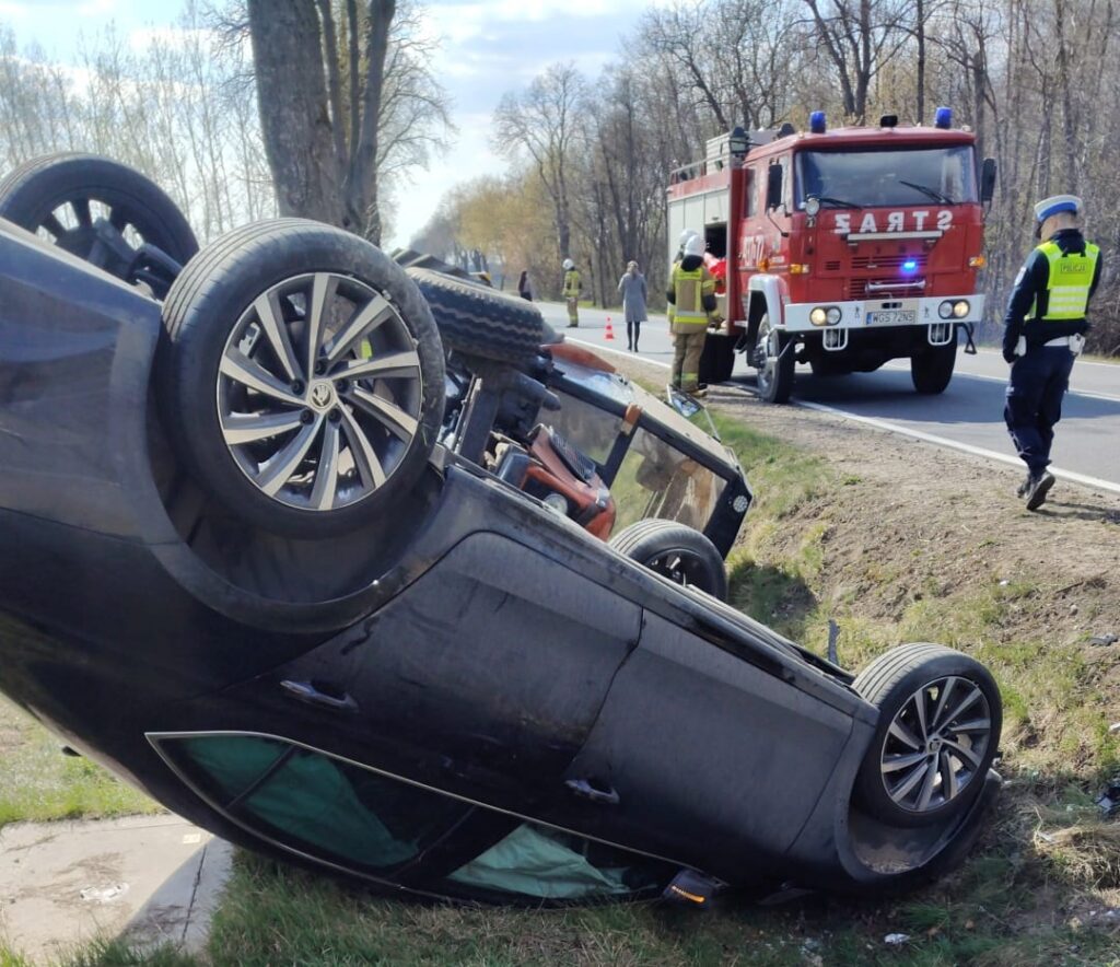 Czarny samochód osobowy leży na dachu przy drodze, obok niego strażacy i policja pracują nad zdarzeniem drogowym.