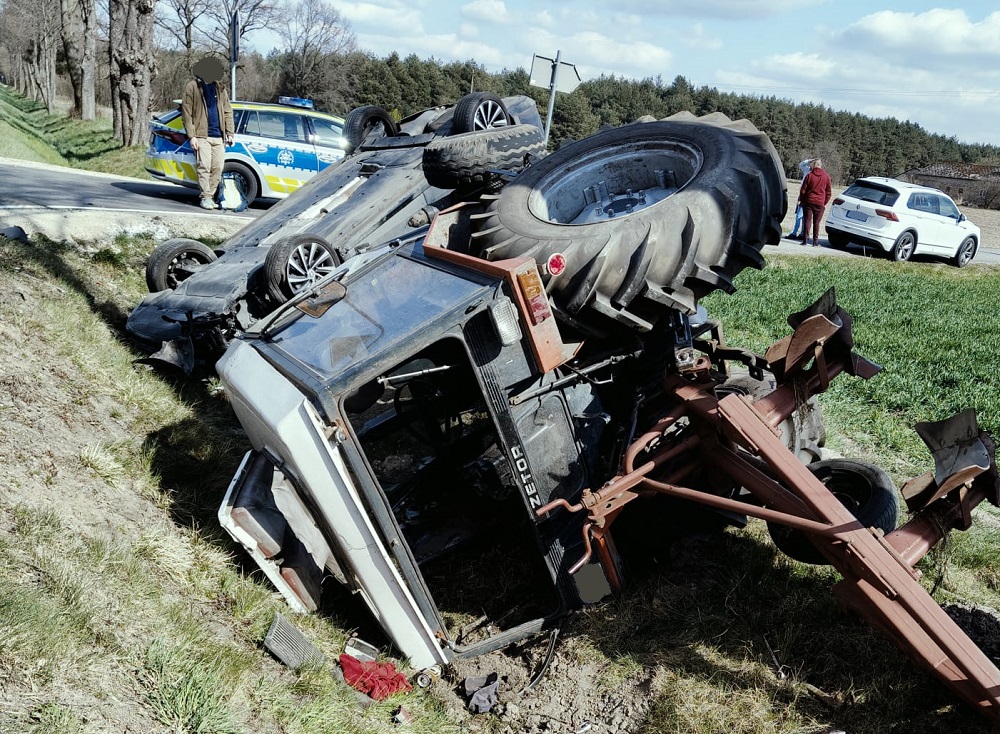 Traktor leży na boku obok drogi, a w tle widać policyjny radiowóz oraz osoby obserwujące zdarzenie.