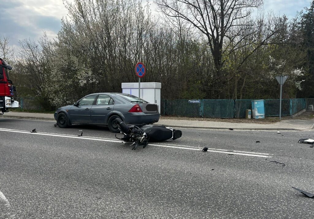 Motocykl leży na jezdni obok szarego samochodu osobowego, w tle znak zakazu parkowania i zarośla.