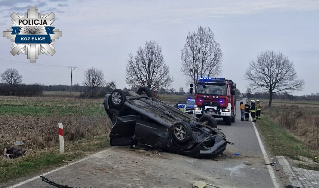 Rozwrócone auto osobowe leży na poboczu drogi, obok którego pracują strażacy i policjanci.