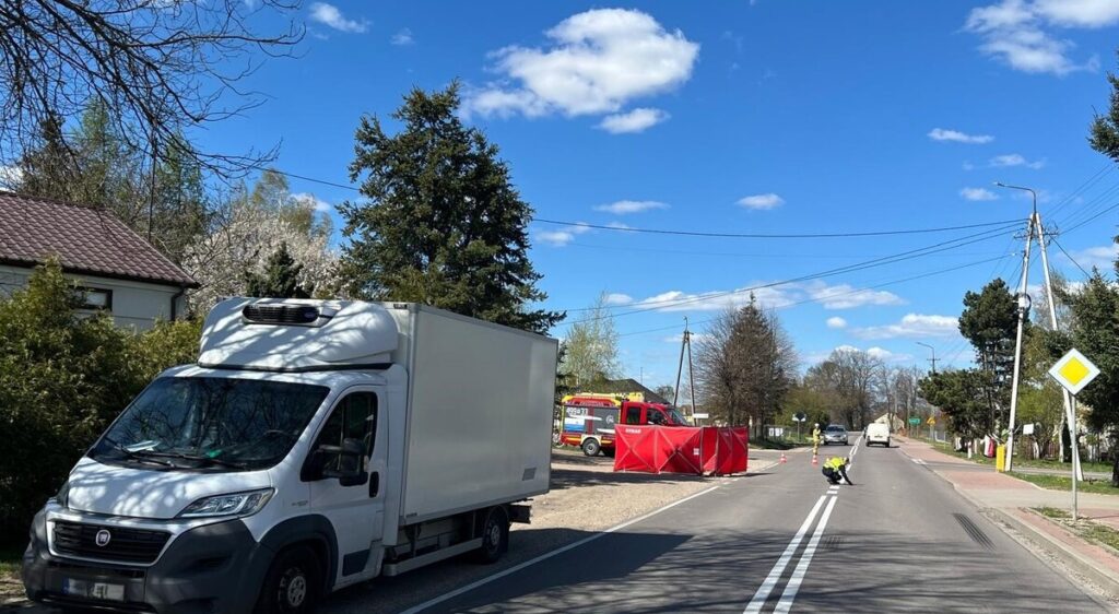 Biały samochód dostawczy zaparkowany przy drodze, obok czerwony wóz strażacki oraz pracownik w kamizelce odblaskowej.
